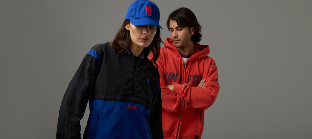 Two people wearing colorful jackets and a blue cap against a gray background
