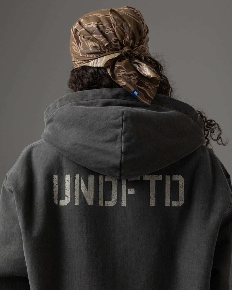 Back view of a person wearing a dark charcoal gray hoodie with 'UNDFTD' printed in distressed light gray letters across the upper back, paired with a brown camouflage headscarf, against a solid gray studio background.