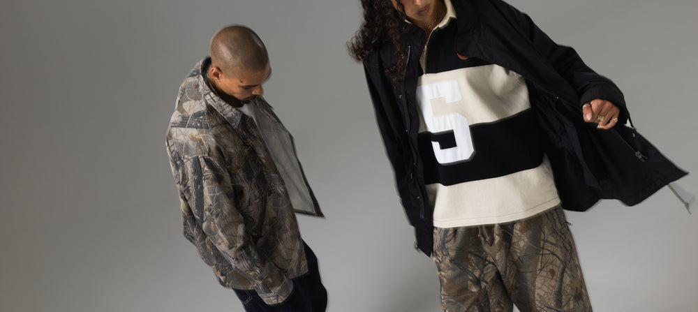 Two models wearing oversized camouflage jackets and matching camo pants, posed against a plain gray studio background.