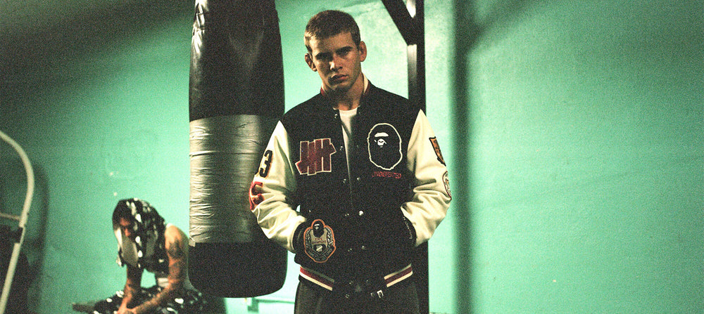 A man wearing a black and cream varsity jacket with A Bathing Ape (BAPE) ape head patches stands in a gym in front of a hanging punching bag. Another person is visible in the background.