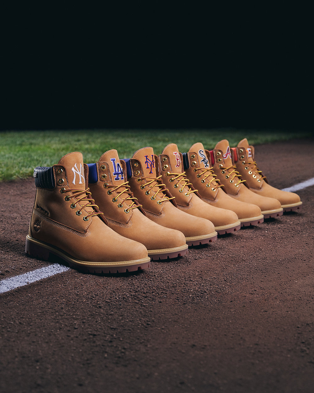A row of tan leather baseball boots lined up on a baseball field dirt, each featuring a different MLB team logo on the tongue, including New York Yankees, Los Angeles Dodgers, San Francisco Giants, Seattle Mariners, Houston Astros, and Chicago Cubs.