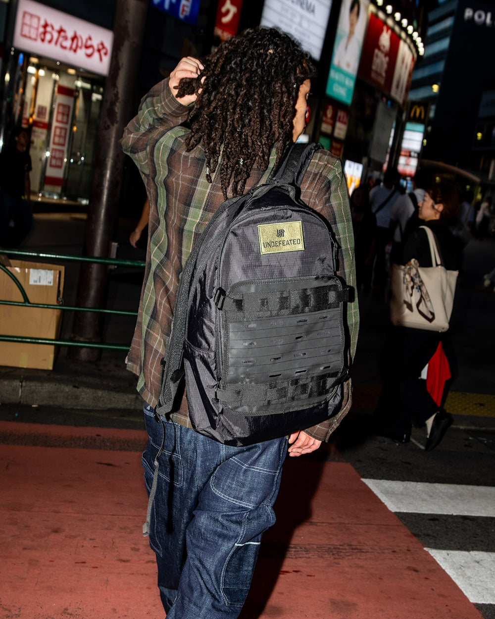 Person with long dreadlocks, seen from behind, walking a city street at night in a plaid shirt and loose jeans, carrying a black backpack labeled UNDEFEATED.
