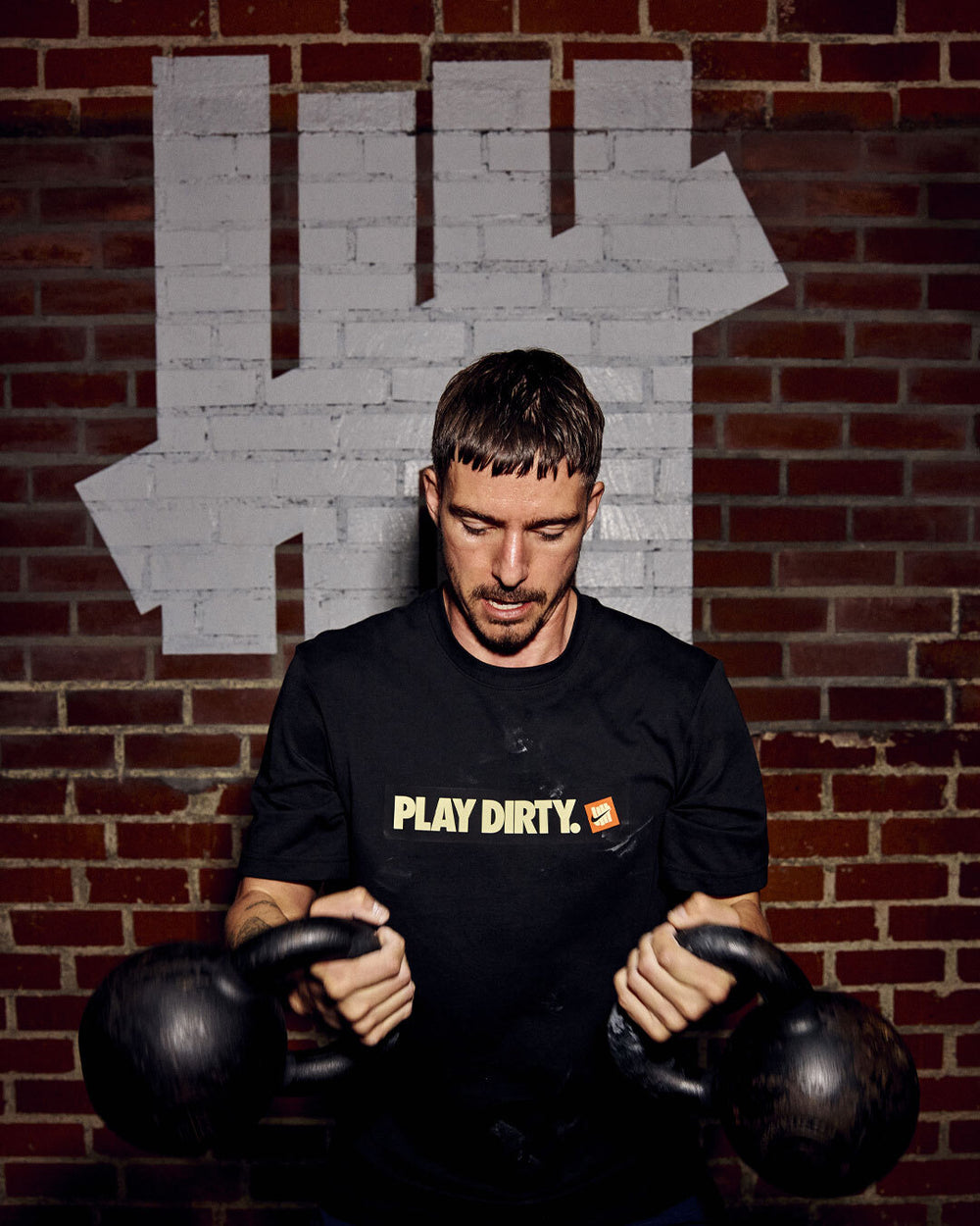 Man holding kettlebells in front of a brick wall with a large white 5 strike on it.
