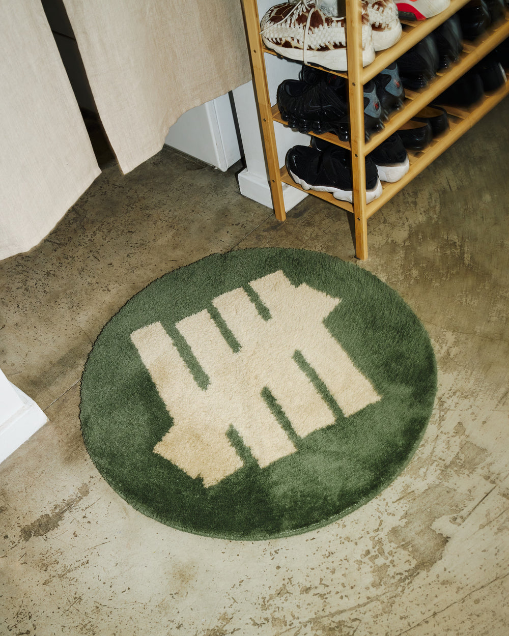 Green rug with a beige five strike logo on a concrete floor next to a wooden shoe rack.