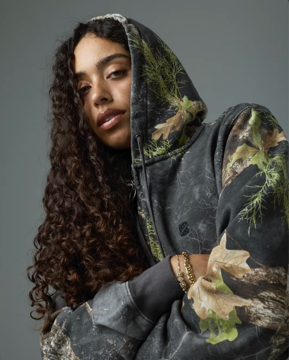 A model wearing a dark gray hooded sweatshirt with a real tree camouflage print, featuring green moss textures and brown leaf details, set against a solid gray studio background.