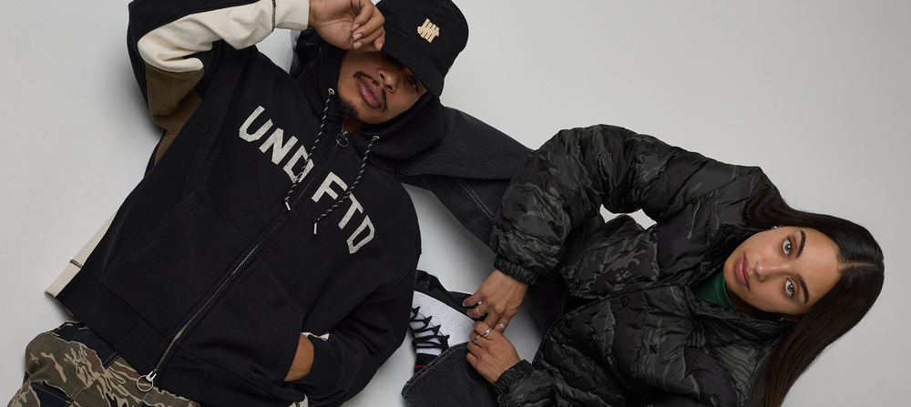 Two models lying on a light gray floor wearing streetwear apparel. The model on the left wears a black zip-up hoodie with 'UNDFTD' printed in white across the chest, a black cap with a gold logo, and camouflage pants.